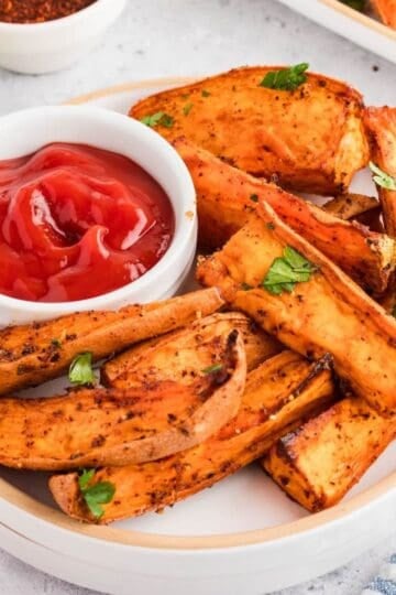 Air fryer sweet potato wedges on a white plate with a small bowl of ketchup.