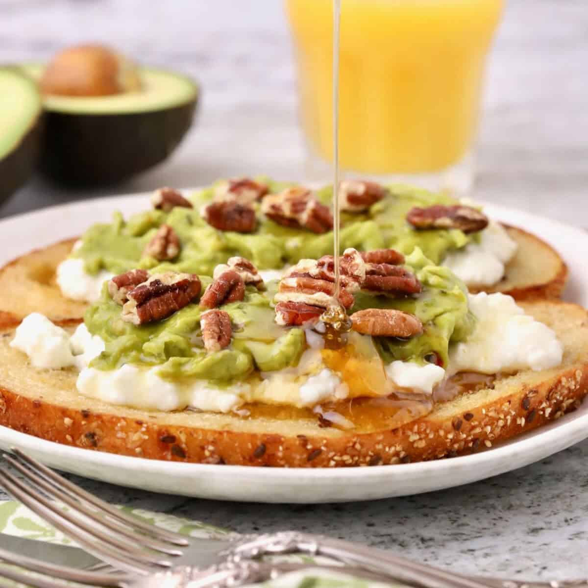 Avocado toast on a plate topped with avocado, cottage cheese and pecans, with a drizzle of honey.