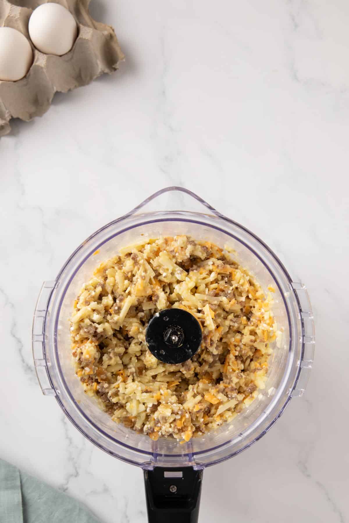Shredded hash browns, cheeses, onions, and sausage are being combined in a food processor. 