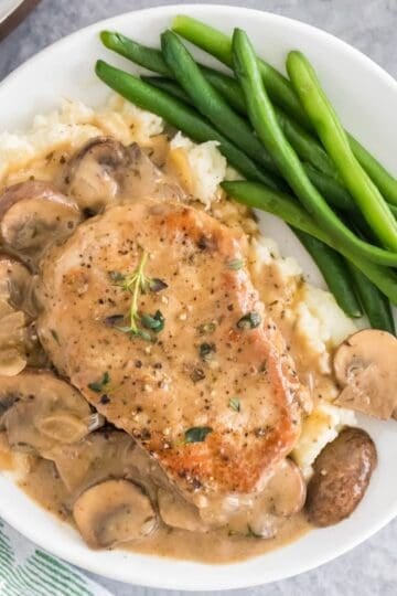 Creamy mushroom pork chops are served on a white plate over rice with mushrooms, sauce, and green beans..