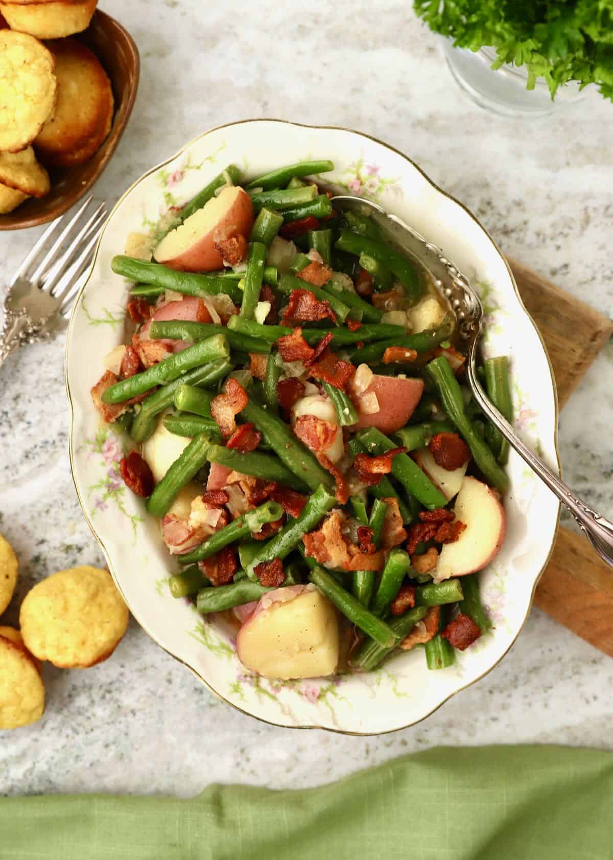 A white bowl with dainty pink flowers is full of cooked green beans and potatoes, and garnished with bacon. 