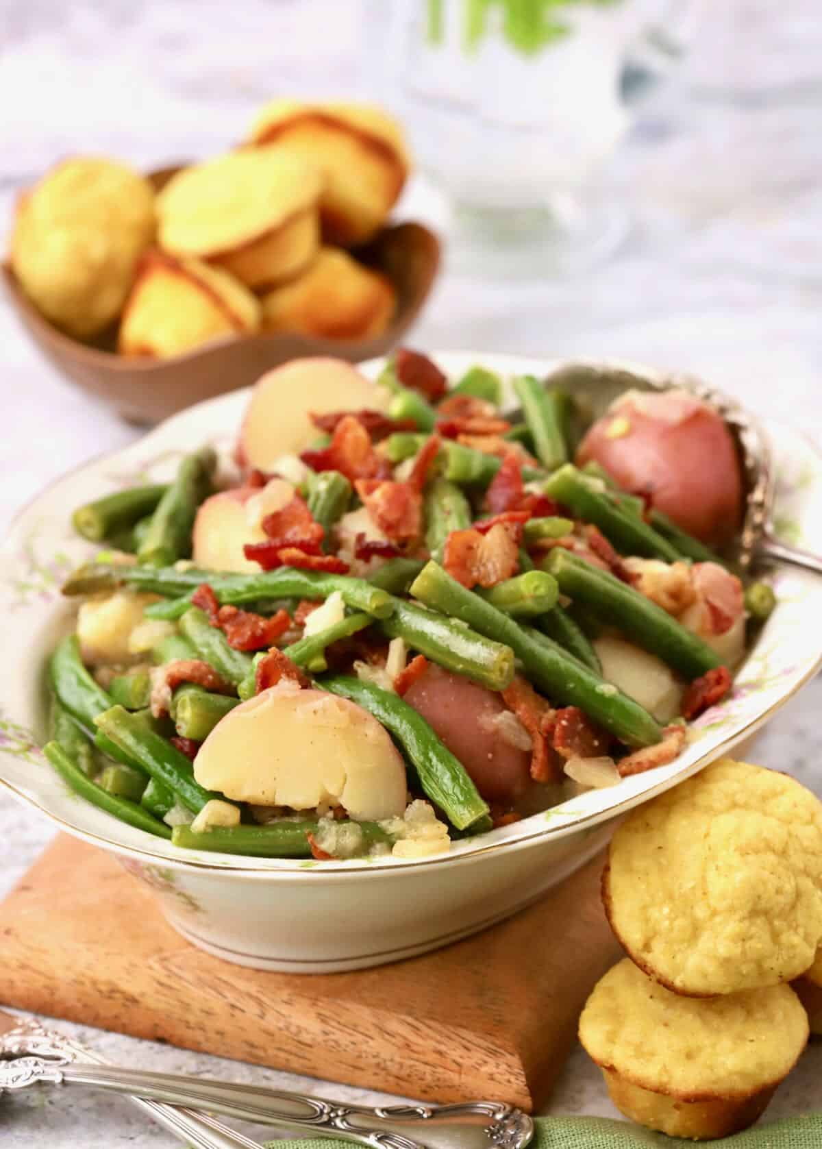 A china serving bowl full of cooked green beans and potatoes garnished with bacon.