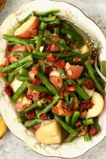 A china bowl is full of cooked green beans and potatoes, and garnished with chopped bacon. Small corn muffins are also present.