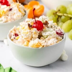 A white bowl full of ambrosia fruit salad with oranges, cherries, grapes, coconut, and pecans, mixed with Cool Whip.