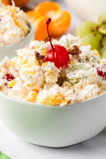 A white bowl full of ambrosia fruit salad with oranges, cherries, grapes, coconut, and pecans, mixed with Cool Whip.