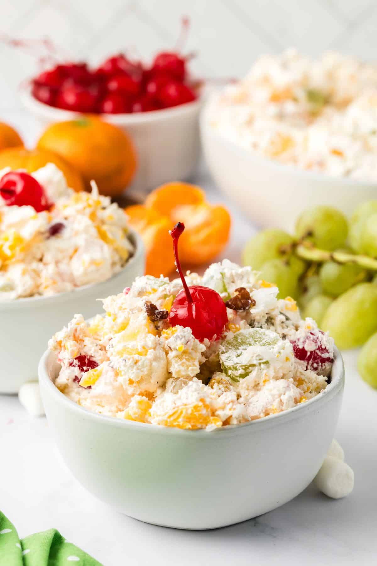 A single serving of ambrosia fruit salad in a small white bowl, garnished with a cherry on top. 