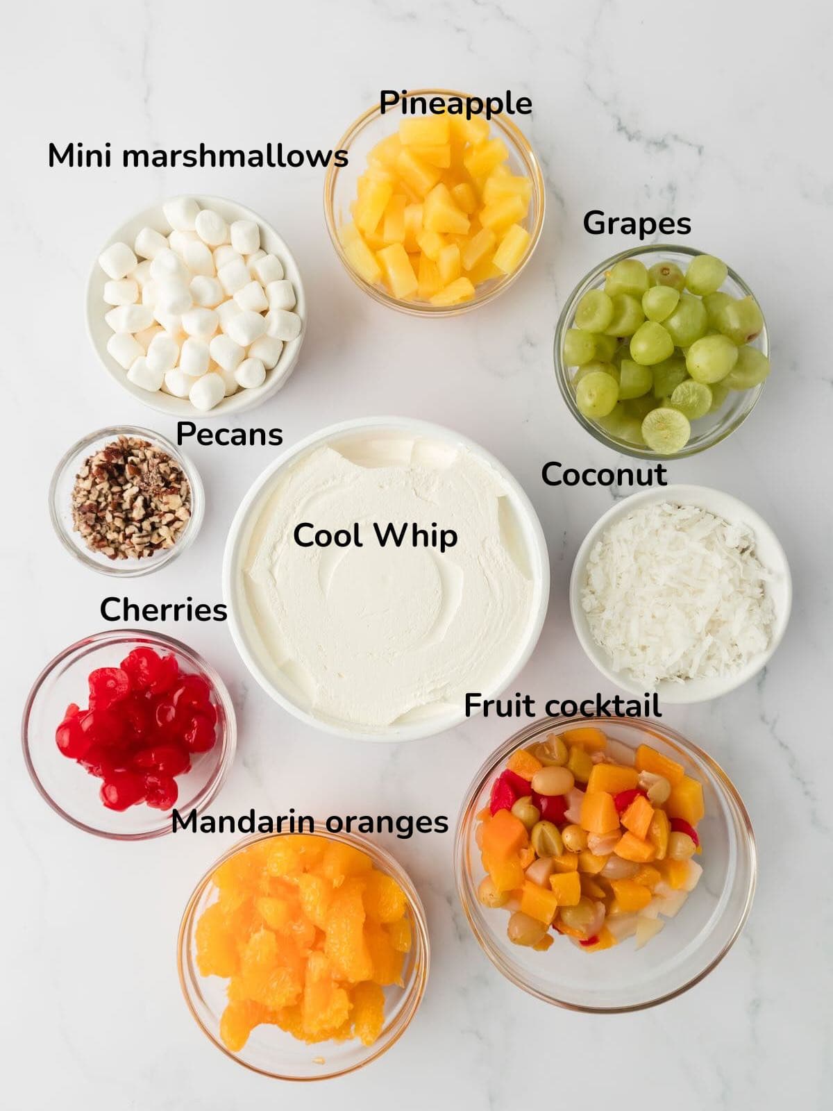 Ingredients to make Ambrosia Fruit Salad include, Cool Whip, oranges, cherries, grapes, fruit cocktail, coconut, and chopped pecans. 