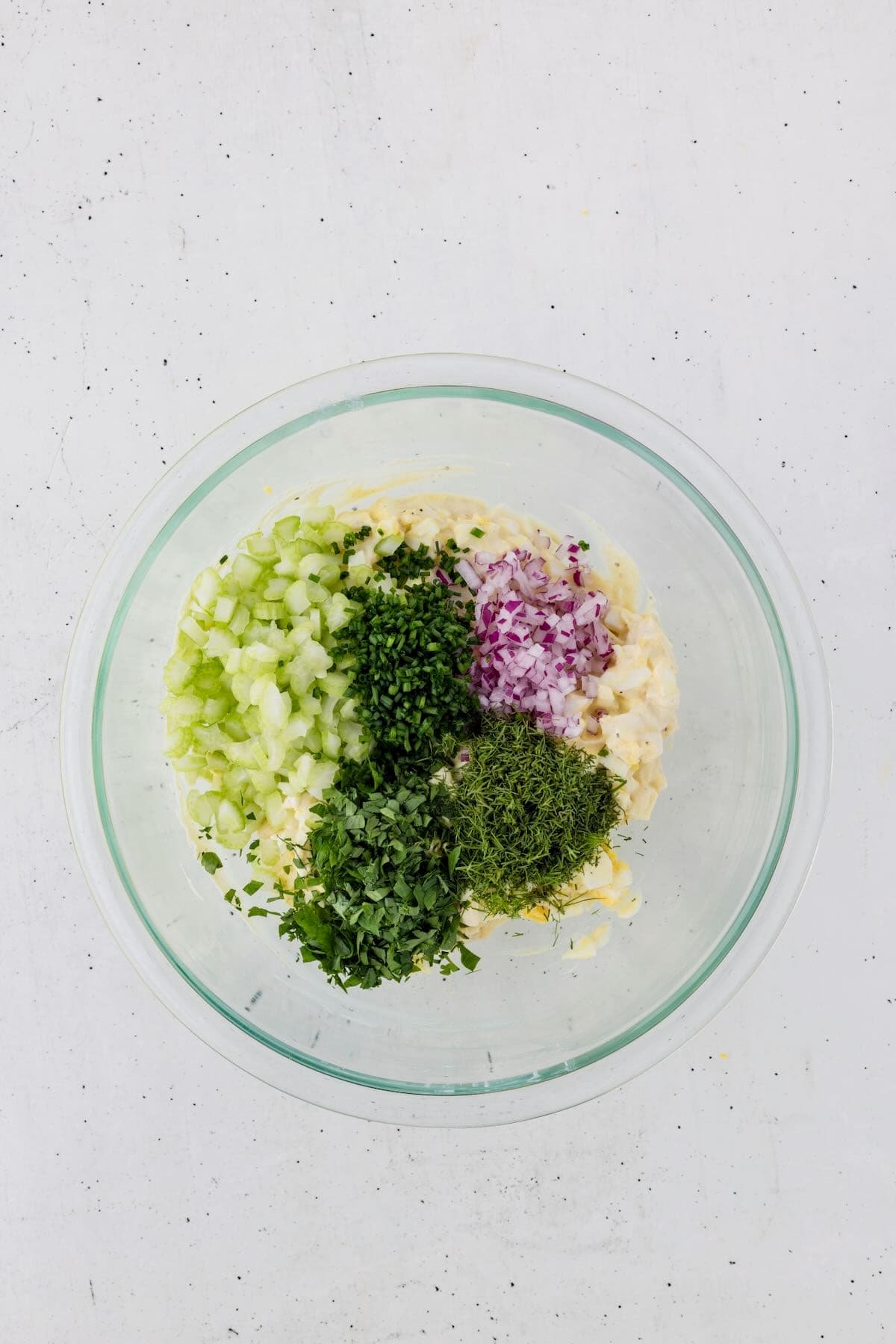 Egg salad in a bowl topped with add-ins including chopped onions, celery, and herbs.