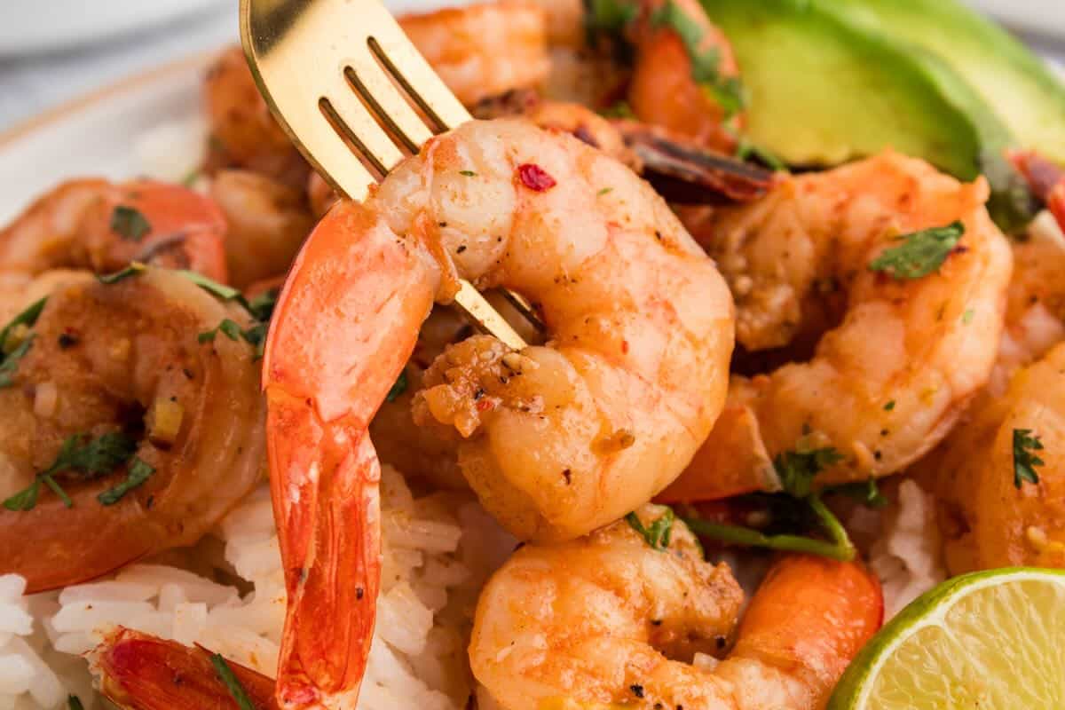 Close-up of tequila lime shrimp on rice with cilantro and lime wedge, pierced with a gold fork for serving.