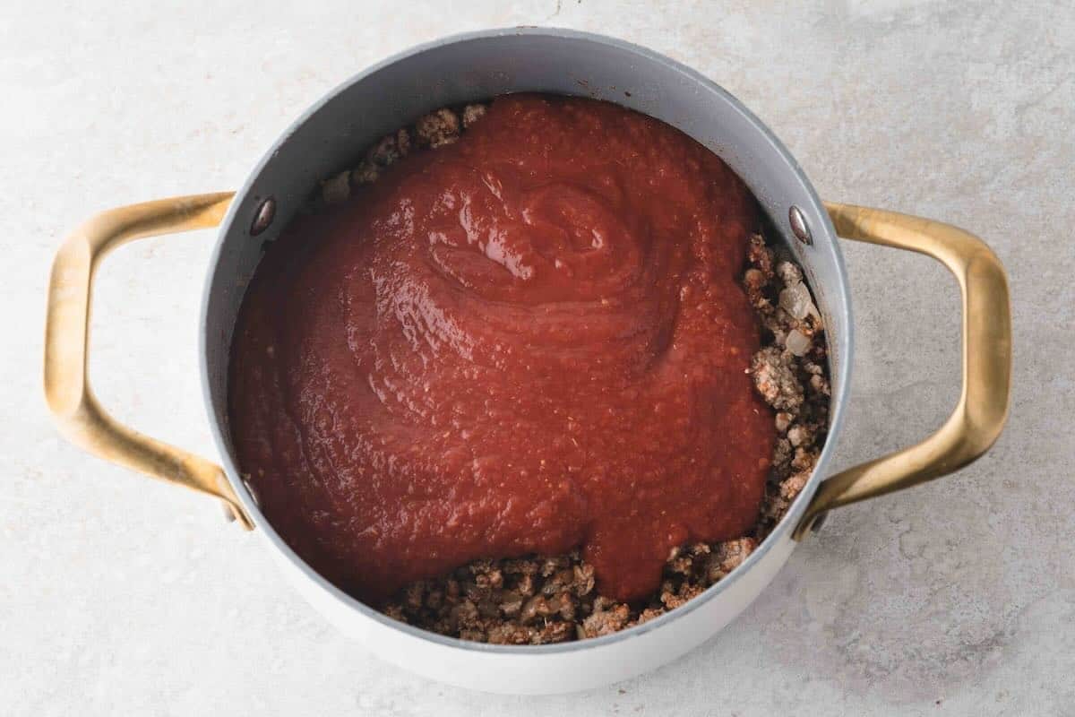 Adding a can of crushed tomatoes to ground beef in a Dutch oven.