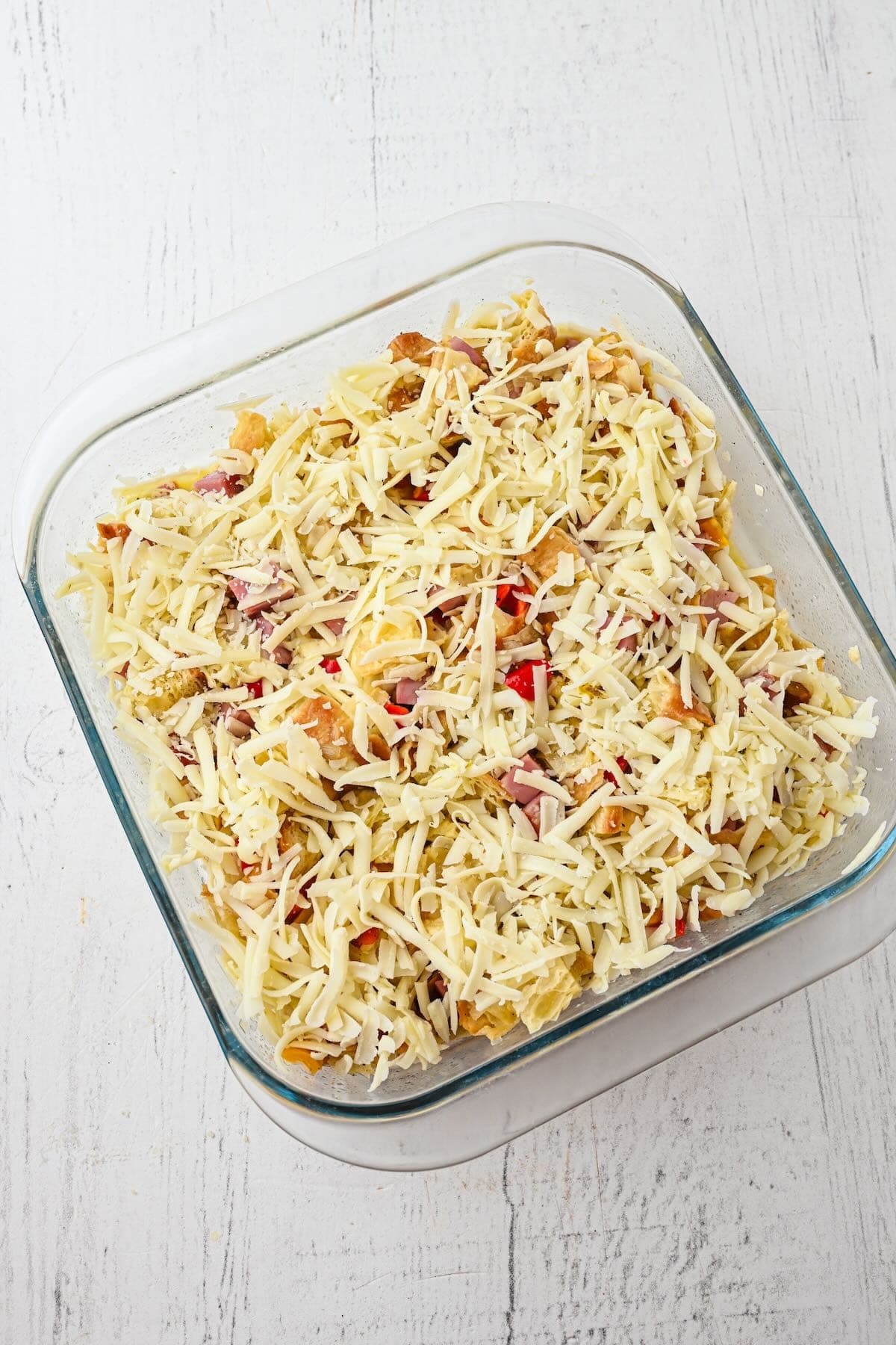 A baking dish full of croissants, ham, peppers, onion, and shredded cheese.