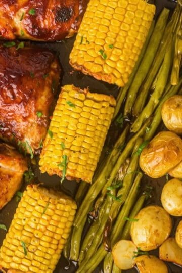A sheet pan dinner including BBQ chicken thighs, corn, green beans, and baby potatoes.
