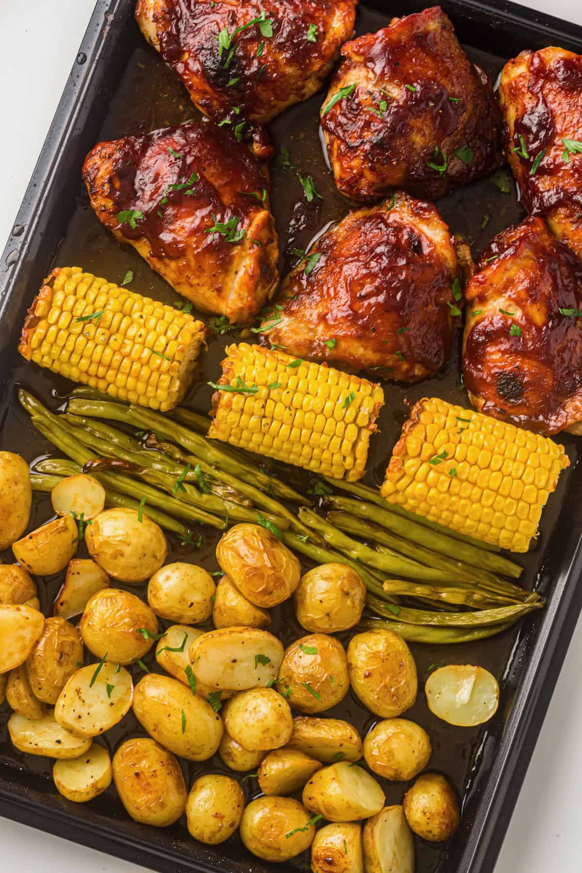 Baked sheet pan BBQ chicken with corn, green beans and halved potatoes garnished with parsley.
