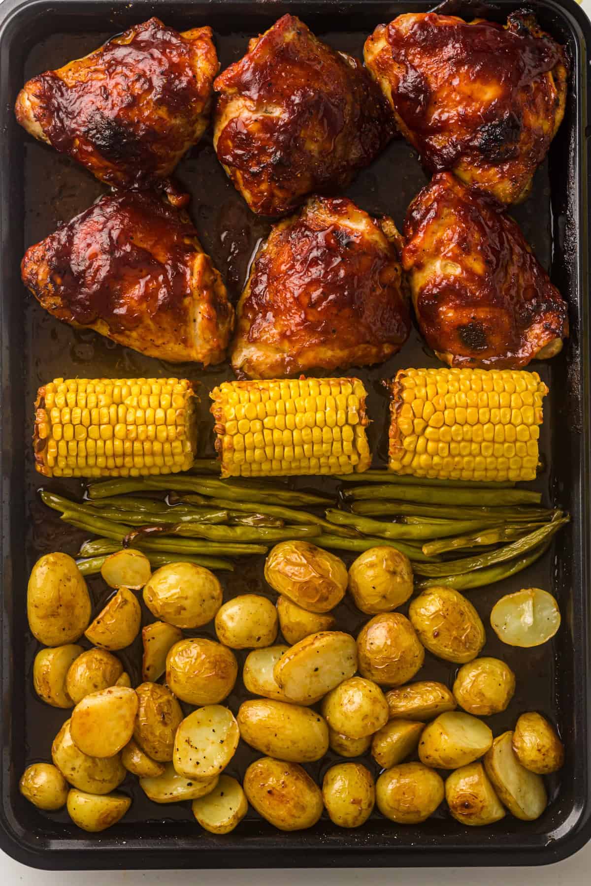 Baked BBQ chicken, green beans, corn, and potatoes on a sheet pan.